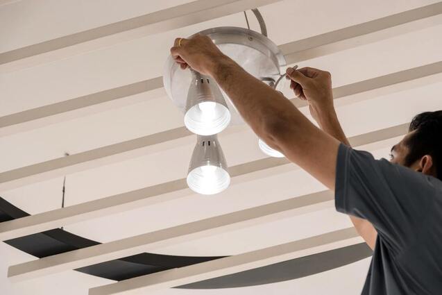 Electrician fitting a set of three lights onto a ceiling with white rafters.