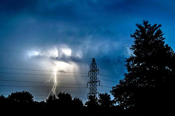 A bolt of lightning in a dark sky with power lines and trees in the foreground.